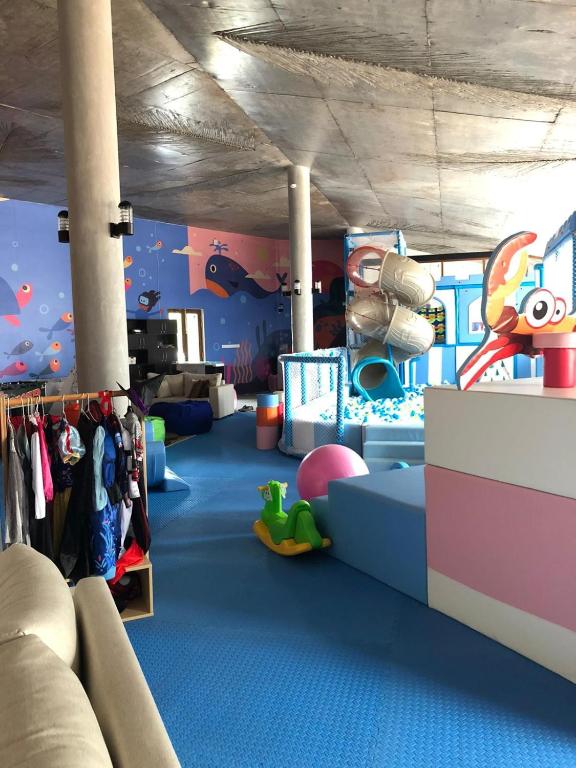 Krāsaina bērnu spēļu istaba ar slidkalniņiem, mīkstām rotaļlietām un jūras motīvu sienām. Telpā redzamas dažādas bērnu drēbes un mīksti sēdekļi.
