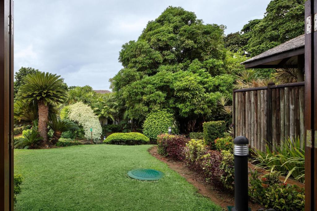 Skats uz zaļu dārzu ar dažādiem augiem un kokiem, kas rada mierīgu un dabisku atmosfēru. Dārzā ir arī apgaismojuma stabi un koka žogs, kas piešķir vietai šarmu.