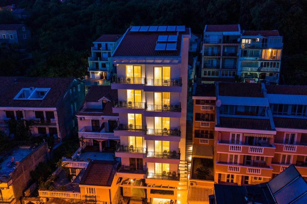 Mūsdienīgs daudzdzīvokļu nams ar apgaismotām terasēm, redzams no augšas, apņemts ar citiem ēkām, kas atrodas kalnainā apvidū. Gaismas spīd caur logiem, radot siltu atmosfēru.