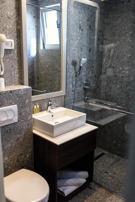 Mūsdienīgs vannas istabas interjers ar akmens apdari, stikla dušu un elegantu izlietni uz koka skapīša. Gaisma ienāk caur logu, radot patīkamu atmosfēru.