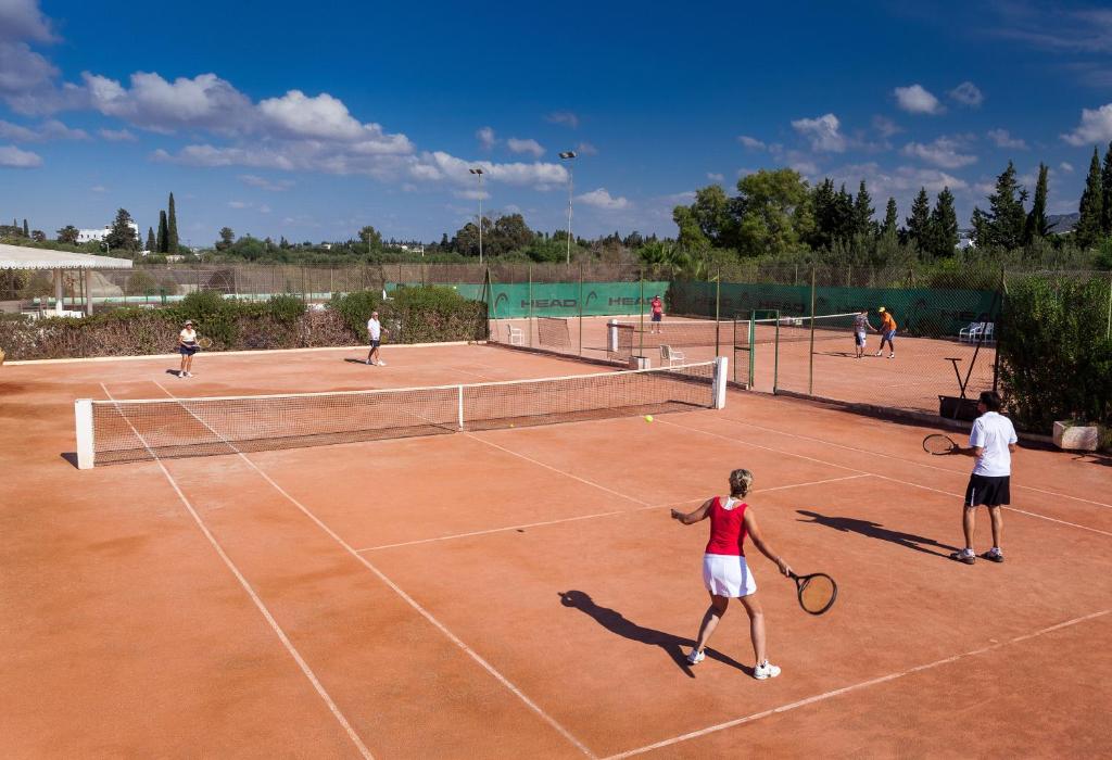 Tennisa laukums ar vairākiem spēlētājiem, kas spēlē dažādās pusēs, un skaidrs debesis fonā.