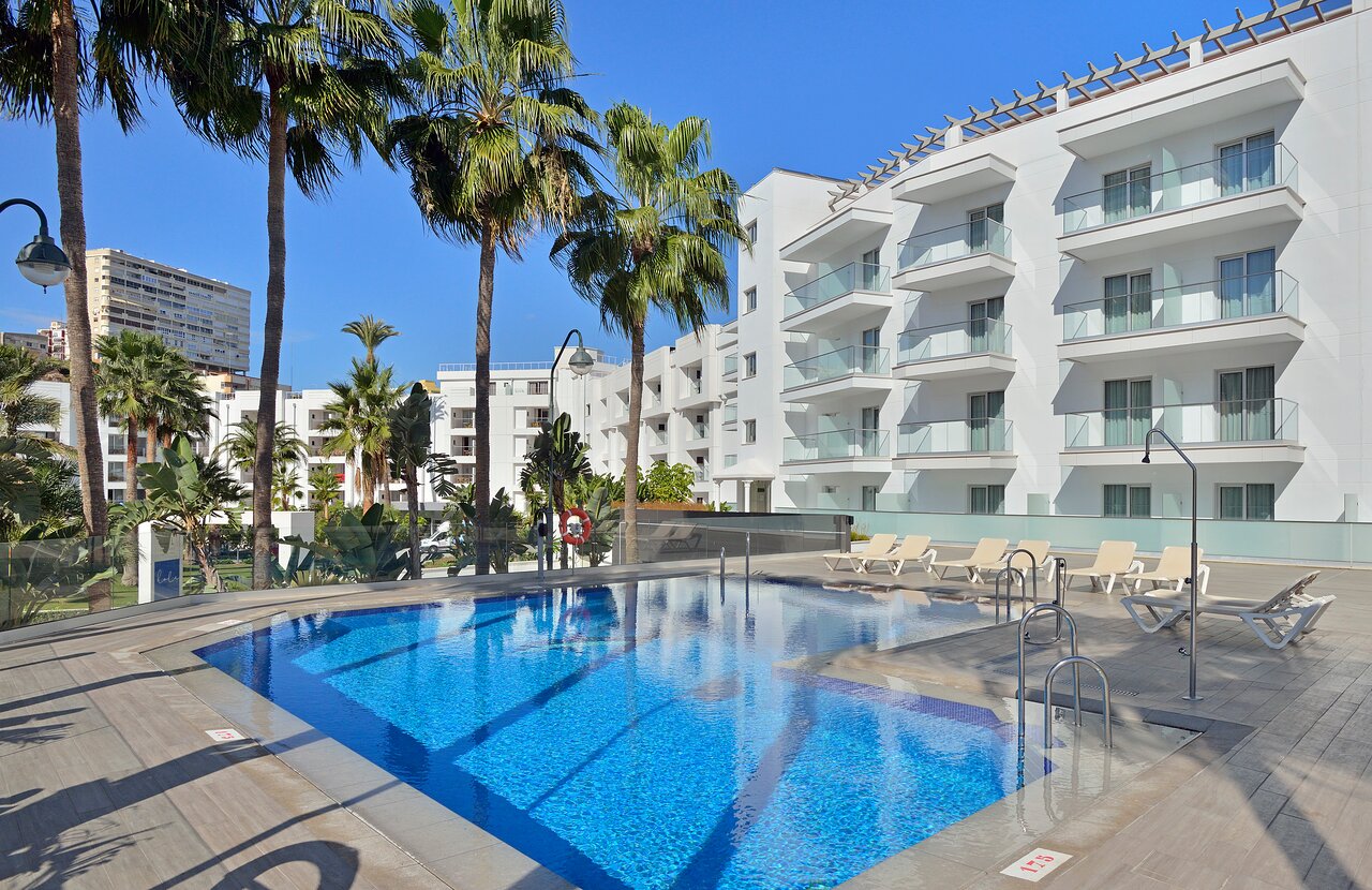 Mūsdienīgs viesnīcas baseins ar palmām un saulessargiem, kas atrodas pie baltas ēkas zem skaidras debess. Ideāla vieta atpūtai un relaksācijai. panoramic view with beach and pools