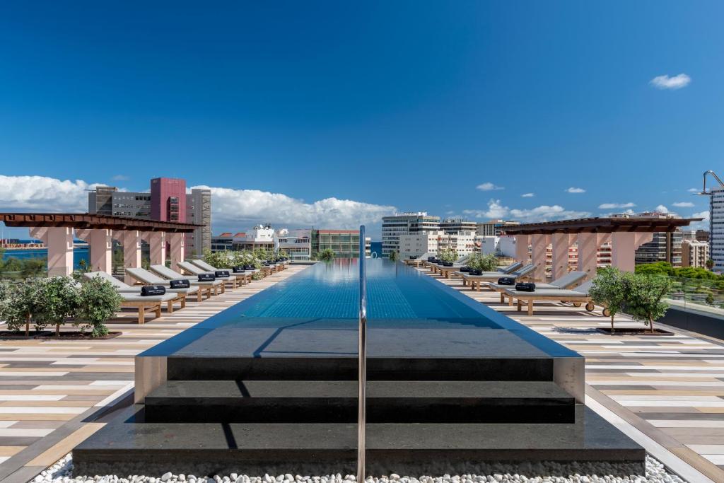 Moderns peldbaseins ar skatu uz pilsētu, apjozts ar sauļošanās krēsliem un zaļiem augiem. Skaidra debesis un mūsdienīga arhitektūra papildina ainavu.