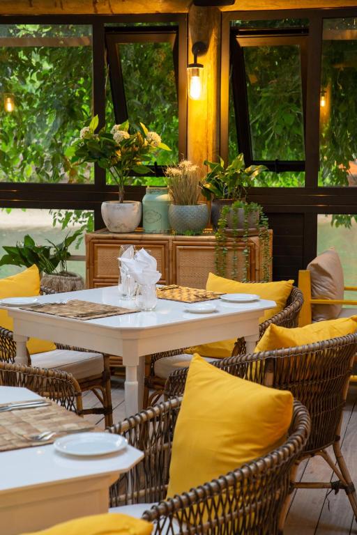 Mūsdienīga restorāna iekārtojums ar baltu galdu, krāsainām spilvenām un zaļiem augiem, kas rada mājīgu atmosfēru. Gaisma un dārza skats papildina patīkamo vidi.