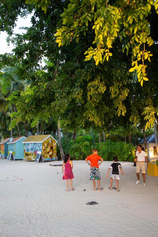 Bērni spēlējas pludmalē, apkārt ir krāsaini namiņi un zaļa daba. Gaisma ir maiga, radot patīkamu atmosfēru.