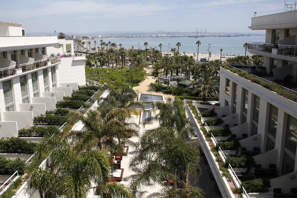 Mūsdienīgs viesnīcas dārzs ar palmu kokiem un skatu uz pludmali, kas izvietota starp ēkām. Fons veido jūra un debesis.