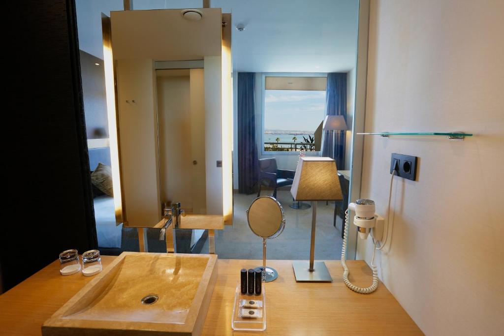 Mūsdienīgs viesnīcas vannas istabas interjers ar skatu uz jūru, ietverot izsmalcinātu izlietni, spoguli un apgaismojumu. Ideāla vide relaksācijai un komfortam.