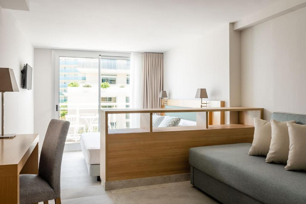 Mūsdienīgs viesnīcas numurs ar gaišu interjeru, ērtu dīvānu un darba galdu, kas piedāvā piekļuvi balkona durvīm. Logā redzams skats uz ārpusi, radot relaksējošu atmosfēru.