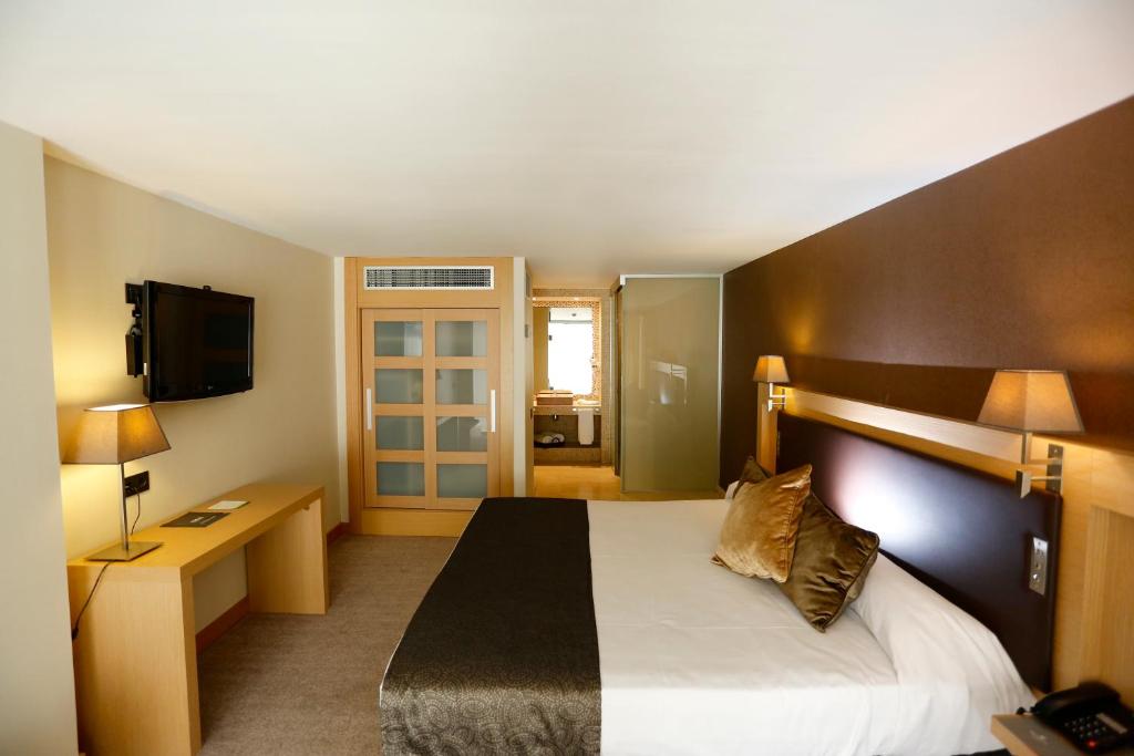 Mūsdienīga viesnīcas istaba ar divguļamo gultu, televizoru un darba galdu, iekārtota ar siltām krāsām un apgaismojumu. Istabā ir stikla durvis, kas ved uz vannas istabu.