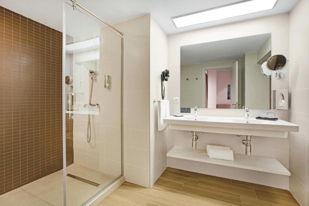 Mūsdienīgs vannas istabas interjers ar stikla dušu, divām izlietnēm un brūnu flīžu akcentu. Telpa ir gaiša un plaša, nodrošinot ērtu un stilīgu vidi.