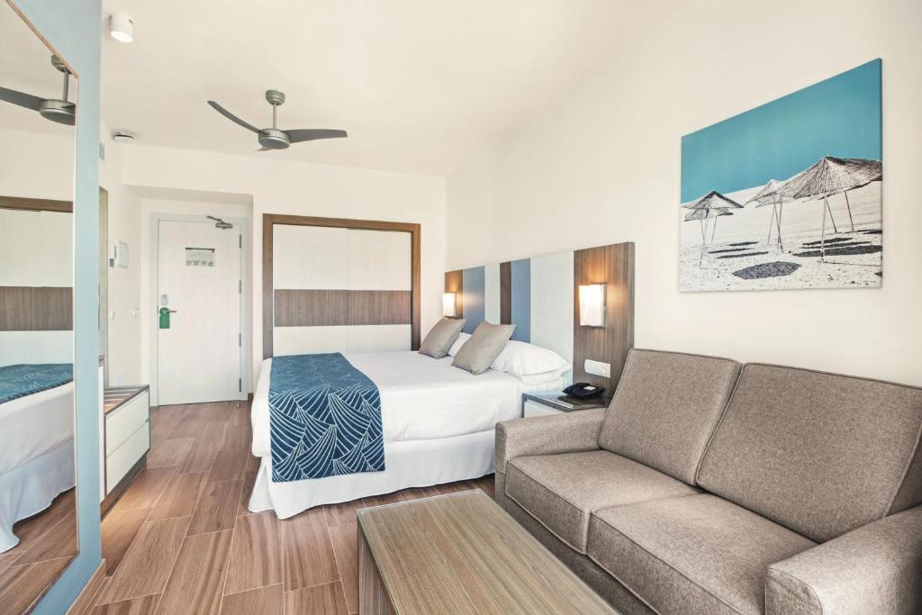 Mūsdienīgs viesnīcas numurs ar divguļamo gultu, dīvānu un plakanā ekrāna televizoru, apgaismots ar siltām gaismām. Uz sienas redzama pludmales ainava.