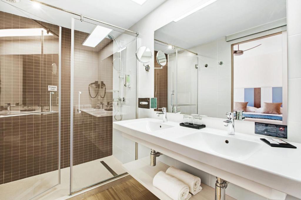 Mūsdienīgs vannas istabas dizains ar dušu un diviem izlietnēm, kas rotā elegantu interjeru ar gaišiem toņiem un koka grīdu.