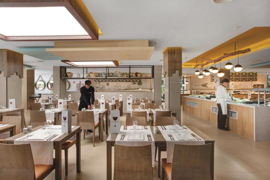 Mūsdienīgas restorāna telpas iekārtojums ar koka mēbelēm un eleganti klātiem galdiem. Apskatāma virtuve un apkalpojošs personāls.