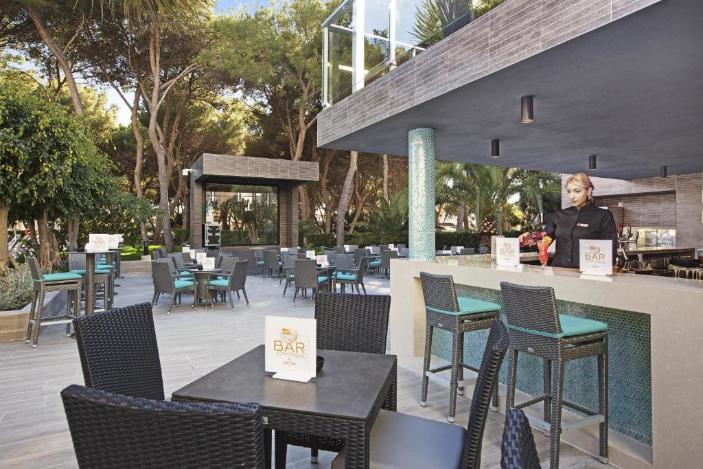 Ārtelpa ar bāru un galdiem, kas izvietoti zem kokiem, radot relaksējošu atmosfēru. Bārmenis gatavo dzērienus, izmantojot modernu bāra aprīkojumu.