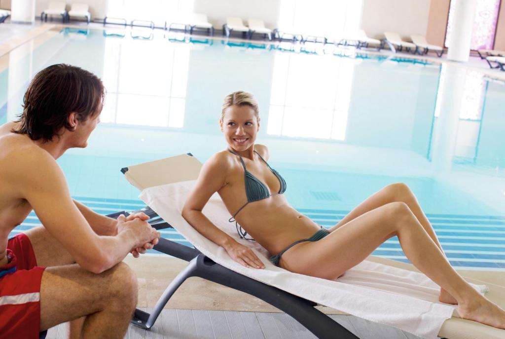 Atpūtas brīdis pie baseina, kur sieviete atpūšas uz sauļošanās krēsla, bet vīrietis ar interesi skatās uz viņu. Fons ir gaišs un ūdens ir dzidrs.