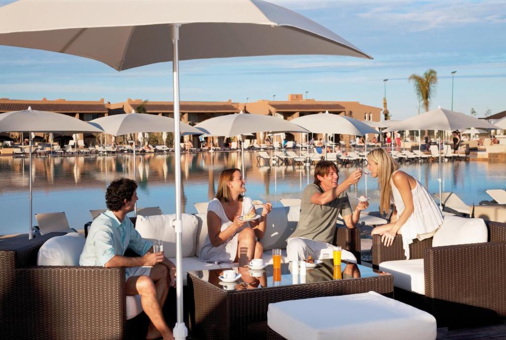 Vasaras atpūta pie baseina, kur draugi bauda dzērienus un uzkodas, sēžot ērtos krēslos zem saulessargiem. Viegla atmosfēra un skaista ainava rada relaksējošu noskaņu.