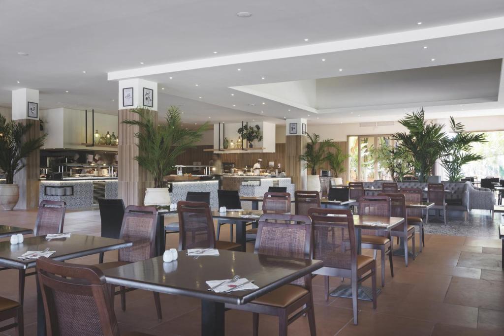 Mūsdienīgs restorāns ar plašu interjeru, kurā ir koka galdi un krēsli, kā arī zaļas palmas. Skats uz atklātu virtuvi un apgaismojumu, kas rada patīkamu atmosfēru.