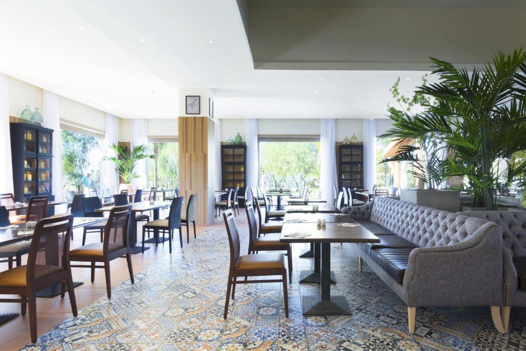 Moderns restorāns ar gaišu interjeru, kas ietver elegantu mēbelējumu un zaļus augus. Plašas logu atveres nodrošina dabīgu apgaismojumu un skatu uz dārzu.