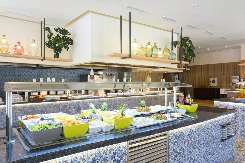 Moderns bufetes stils restorāns ar dažādiem ēdieniem un zaļiem augiem dekorācijās. Redzami šefpavāri, kas gatavo ēdienus.