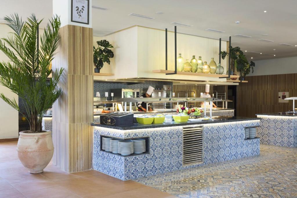 Mūsdienīgs un elegants restorāna interjers ar plašu bufetes zonu, dekorētu ar flīzēm un zaļām augu kompozīcijām, kas rada patīkamu atmosfēru.