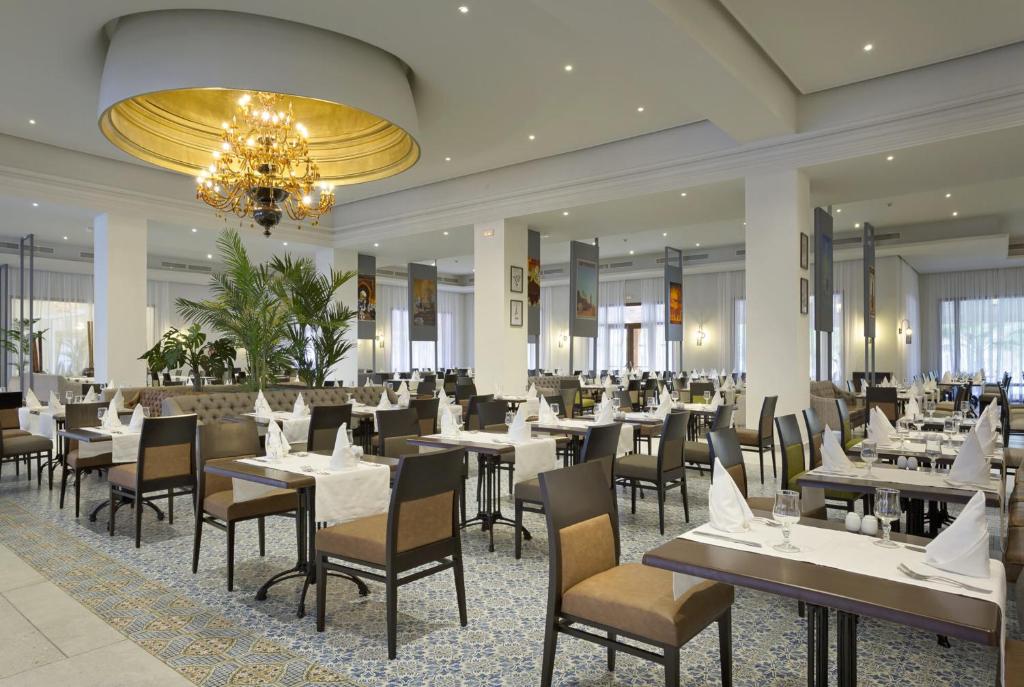 Elegants restorāns ar gaumīgi noformētām galdiņiem, kas rotāti ar baltām galdautām un dekoratīvām servēšanas detaļām. Telpā ir plaša, gaiša atmosfēra ar greznu lustru un augiem, radot patīkamu ēdināšanas pieredzi.