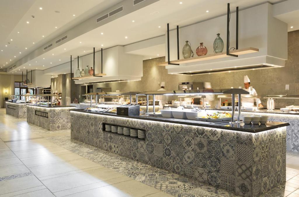 Mūsdienīgs bufetes stils ar dažādiem ēdieniem un šefpavāriem, kas strādā virtuvē. Interjers ir elegants ar stilizētu flīžu apdari un mūsdienīgu apgaismojumu.