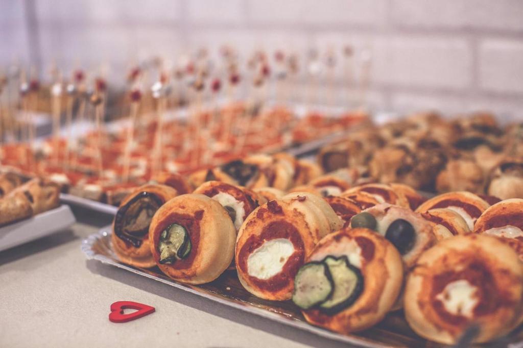 Dažādas uzkodas, tostarp picas rullīši ar dārzeņiem un siera pildījumu, izkārtotas uz galda. Uzkodas rotā palīgi, kas atrodas aizmugurē.