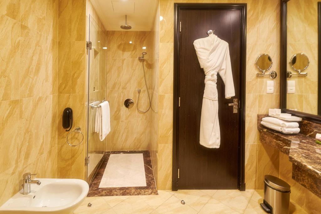 Mūsdienīga vannas istaba ar siltu krāsu paleti, stikla dušu un dvieli, kas pakārts pie durvīm. Vannas istabā ir arī izlietne un spoguli, radot elegantu un funkcionālu telpu.