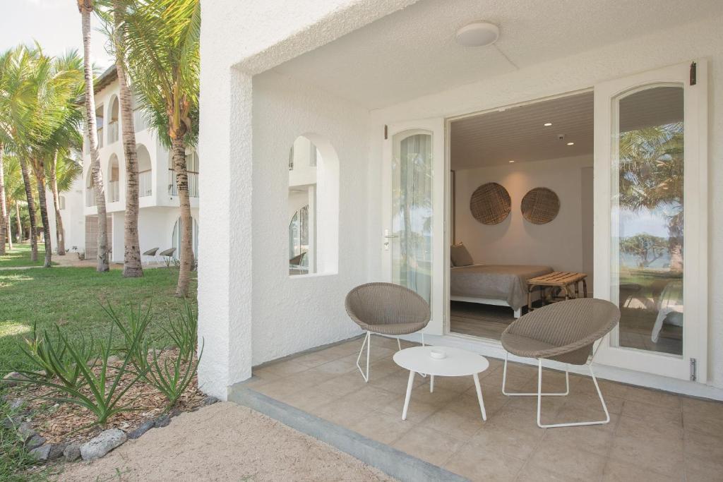 Mūsdienīgs viesnīcas numura balkons ar diviem krēsliem un galdiņu, kas atrodas pie zaļojoša dārza ar palmām. Iekštelpās redzama gulta un dekoratīvi elementi.