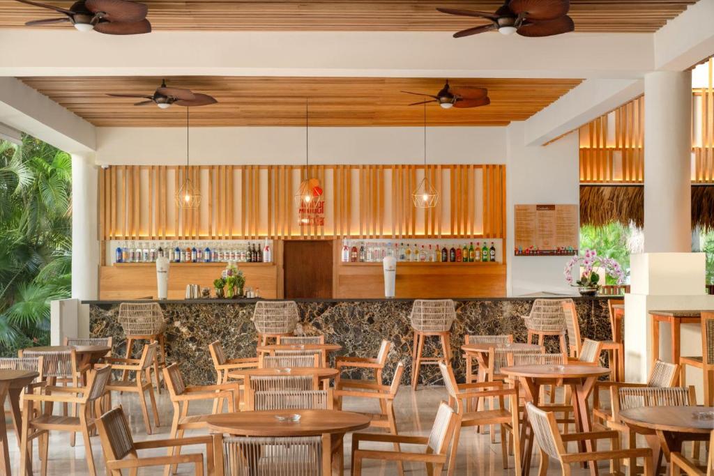 Mūsdienīgs bārs ar koka akcentiem un tropisku zaļumu, kas rada relaksējošu atmosfēru. Kafijas un dzērienu izvēle pie bāra, ap kuriem izvietoti eleganti koka galdi un krēsli.
