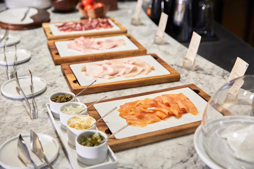 Svaigas gaļas šķēles un garnējumi uz galda, kas izkārtoti uz koka paliktņiem un baltām plāksnēm. Ideāli piemērots brokastīm vai uzkodām.