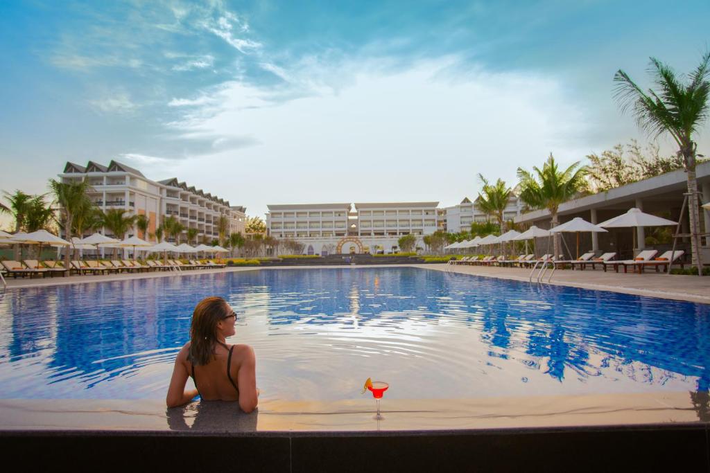 Relaksējoša aina pie viesnīcas baseina ar sievieti, kas baudīja kokteili, un skaistu ainavu ar palmām un viesnīcu ēkām fonā. Ideāla vieta atpūtai un brīvdienām.