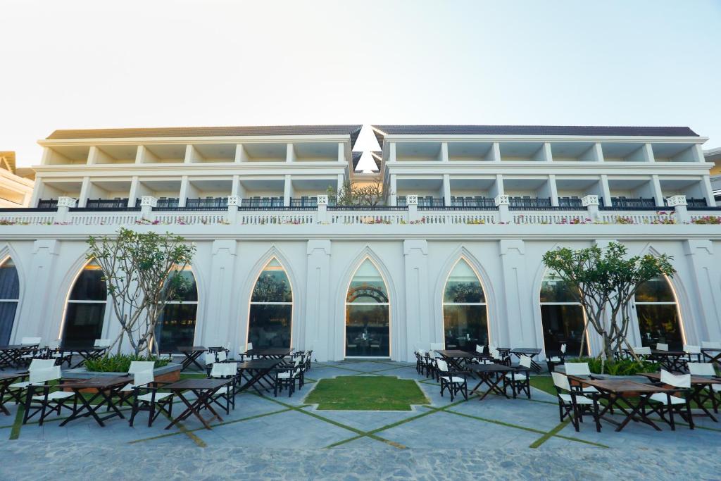 Eleganta viesnīca ar baltām arkveida logiem un plašām terasēm, apkārt esošajiem galdiem un krēsliem. Ideāla vieta relaksācijai un baudīšanai.