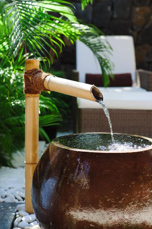 Bambusa strūklaka, kas plūst ūdeni no apaļas podiņa, ieskauta zaļajos augos un atpūtas zonā.