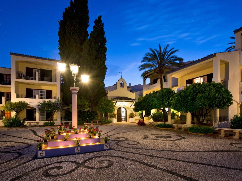 Mierīga viesnīcas iekšpagalma aina ar apgaismotām lampām un ziedu rotājumiem, kas veido pievilcīgu atmosfēru. Apkārt ir zaļi koki un ēkas ar balkoniem, kas piešķir šarmu un relaksējošu noskaņu.