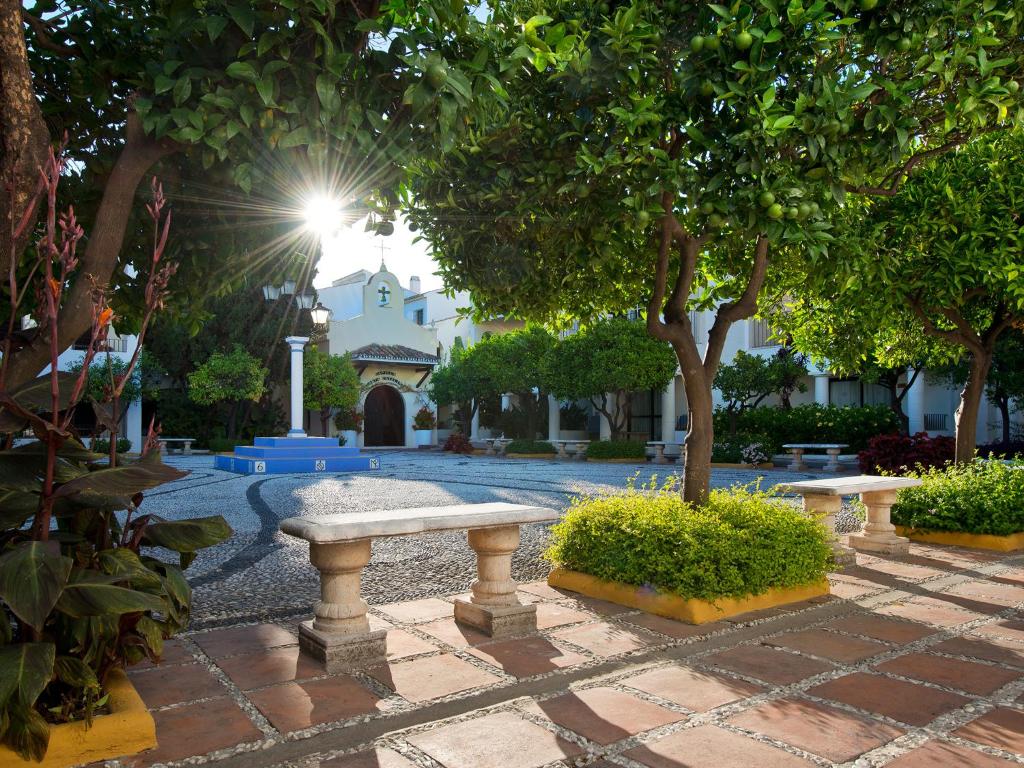 Skaista dārza ainava ar saules gaismu, kas spīd cauri kokiem, akmens soliņiem un ziedošām krūmiem. Mierīga atmosfēra, kas aicina uz relaksāciju un nesteidzīgu atpūtu.