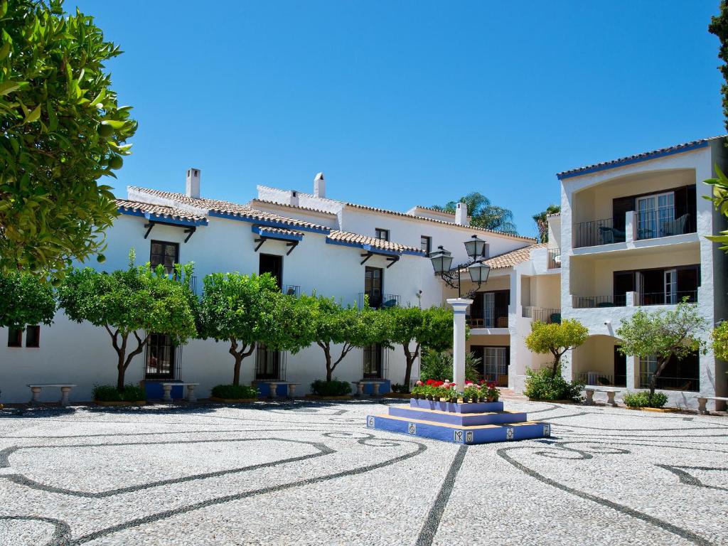 Skaists viesnīcas iekšpagalms ar baltiem ēkām, zaļām kokiem un puķu dobi centrā. Gaisīgs un saulains laiks, kas rada relaksējošu atmosfēru.