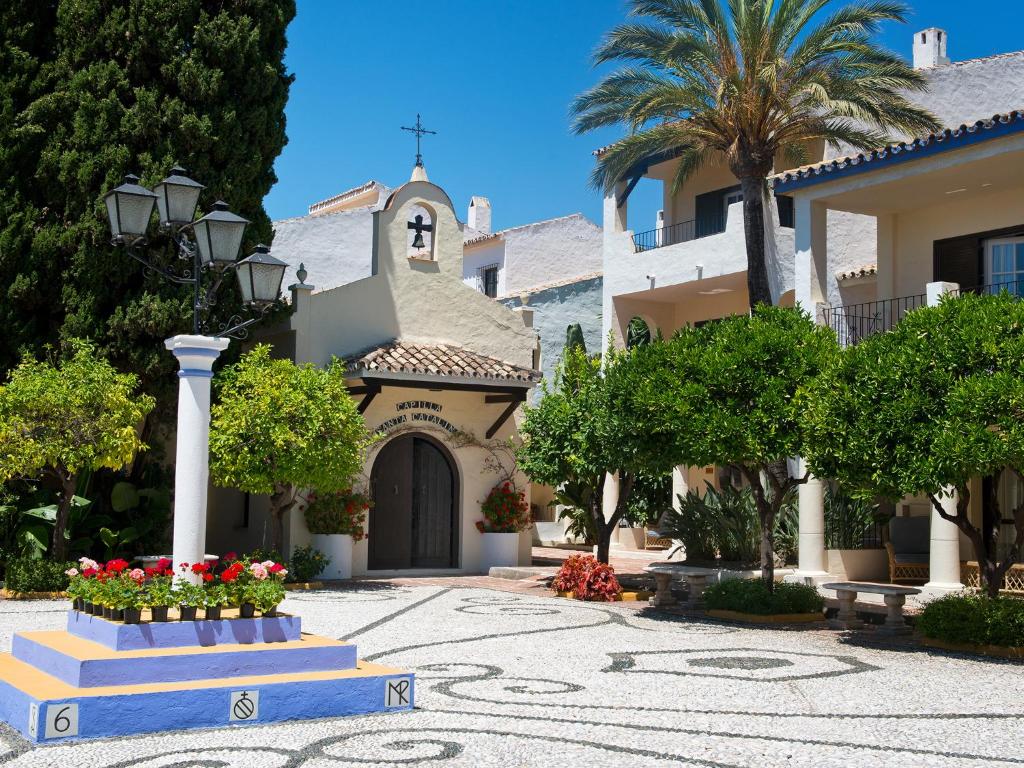 Skaista vidusjūras ainava ar elegantu arhitektūru, ziediem un palmu kokiem, kas veido mierīgu un saulainu atmosfēru. Pilsētas laukums ar dekoratīvu puķu dobi un klasisku lampu.