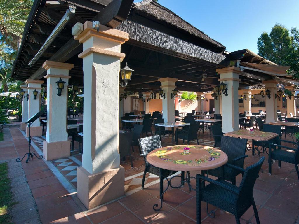 Āra restorāna zona ar elegantiem melniem krēsliem un galdiem, kas dekorēti ar ziediem, siltā saulē. Viegla, atvērtā struktūra ar kolonām un jumtu.