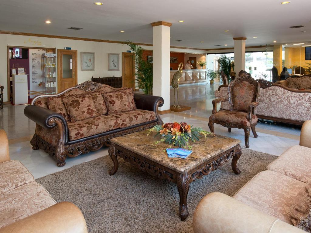 Elegants viesnīcas interjers ar greznām mēbelēm, ieskaitot izsmalcinātu dīvānu un atpūtas krēslu, kas izvietoti ap masīvu galdu. Telpā ir gaiša apgaismojuma un augu akcenti.