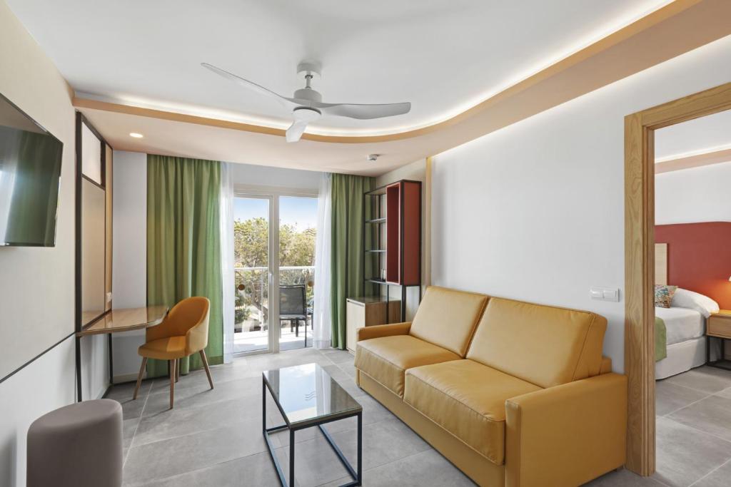 Mūsdienīga viesnīcas numura iekšskats ar dzeltenu dīvānu, rakstāmgaldu un lieliem logiem, kas ved uz balkonu. Interjerā dominē zaļas aizkaru un siltu toņu akcenti.