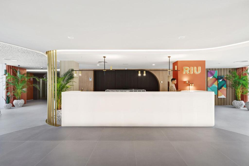 Mūsdienīgs viesnīcas reģistratūras interjers ar elegantu baltu letīti, zelta akcentiem un dzīviem augiem. Aiz letes redzams viesmīlis un krāsaini mākslas darbi uz sienām.