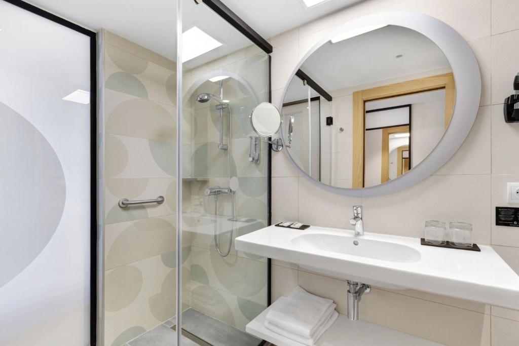 Mūsdienīgs vannas istabas interjers ar stikla dušu, apaļu spoguli un elegantu izlietni. Gaišas krāsas un moderns dizains rada patīkamu atmosfēru.
