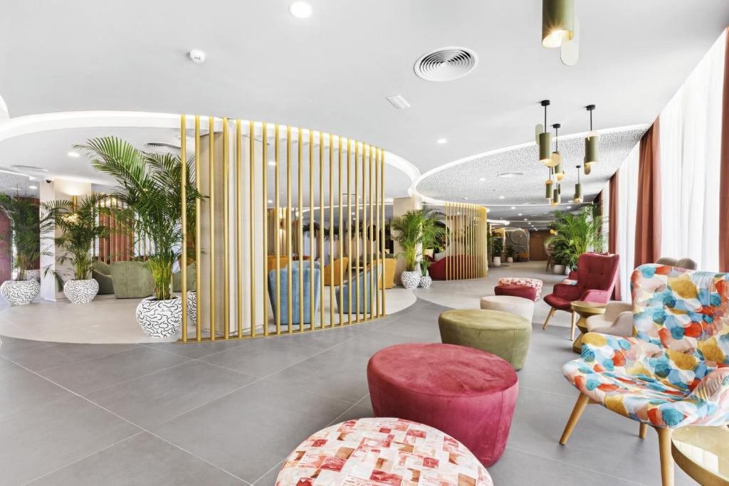 Mūsdienīgs interjers ar krāsainiem krēsliem un dekoratīvām augiem, radot patīkamu un viesmīlīgu atmosfēru. Gaismas un telpas plūsma piešķir dizainam modernu pievilcību.