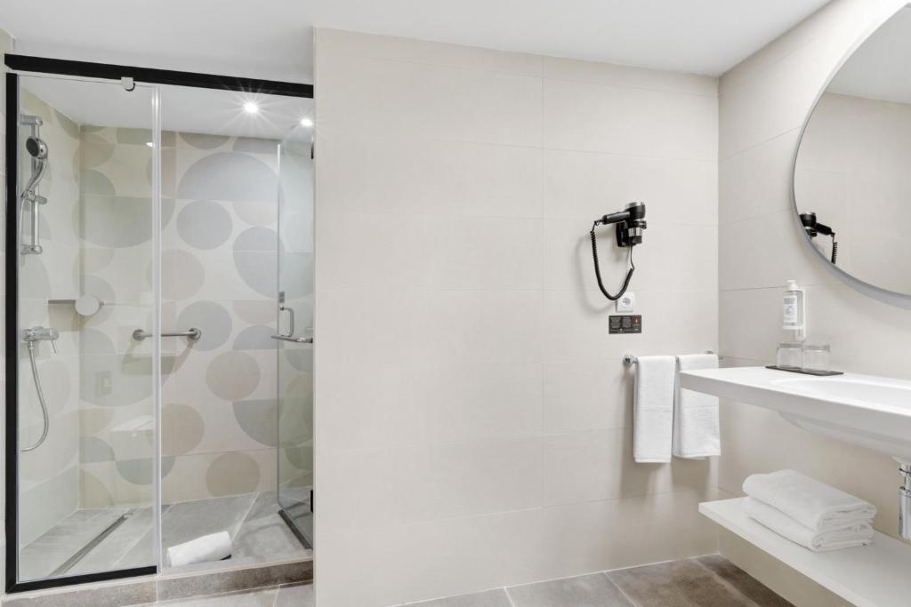 Moderns vannas istabas dizains ar caurspīdīgu dušas kabīni un elegantu spoguli. Telpa ir gaiša un minimālistiska, papildināta ar baltām dvieļiem un higiēnas līdzekļiem.