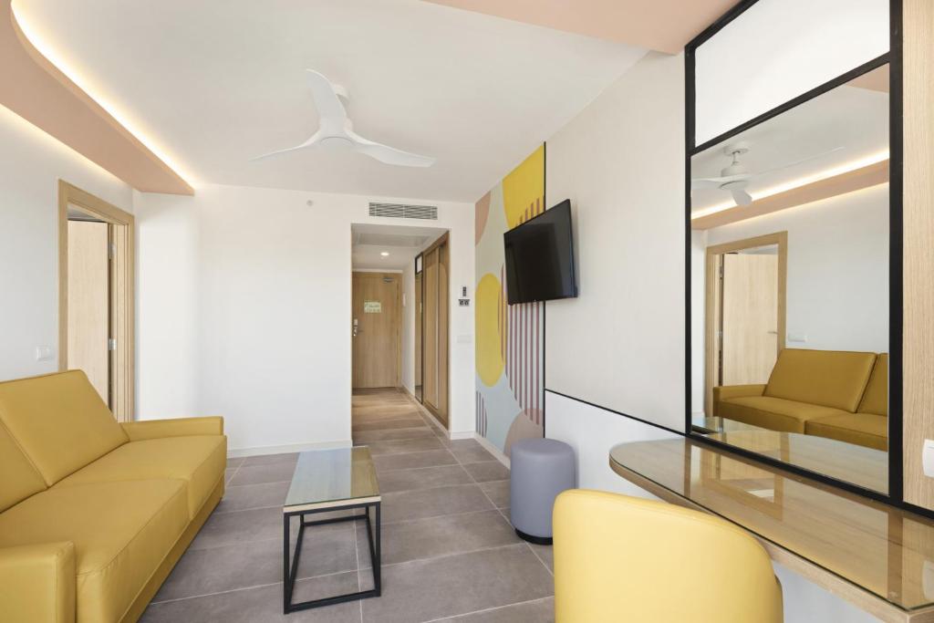 Mūsdienīgs viesnīcas numurs ar dzeltenu dīvānu, stikla galdiņu un modernu interjeru. Telpa ir gaiša ar dekoratīvām sienām un iekārtota ērtai atpūtai.