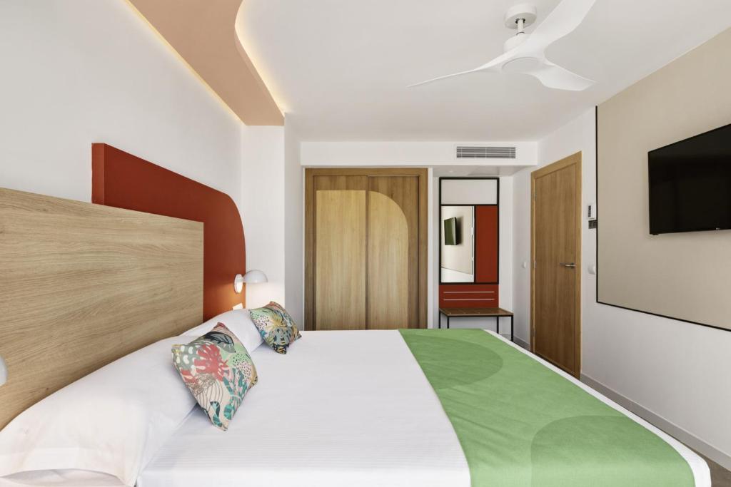 Mūsdienīga viesnīcas numura iekārtojums ar koka gultas galvu, spilveniem ar krāsainiem rakstiem un zaļo gultas veļu. Interjerā ir moderns dizains ar tv un gaismas elementiem.