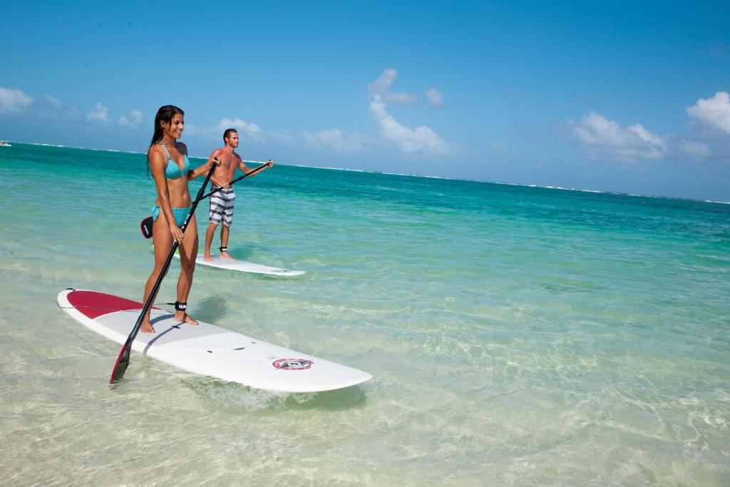 Jauniem cilvēkiem, kas stāv uz sērfošanas dēļiem, baudot sauli un jūras viļņus. Ūdens ir skaidrs un zilgans, ar baltām mākoņiem debesīs.