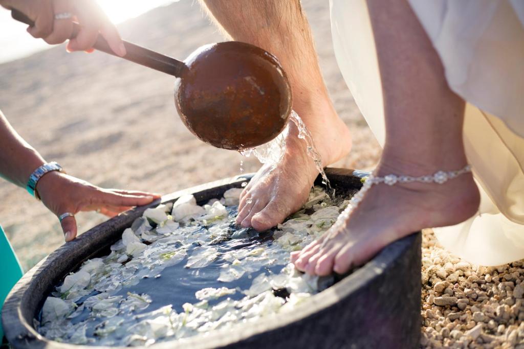 Kāju mazgāšanas ceremonija ar ziedlapiņām ūdenī, kurā divi cilvēki iegremdē kājas, kamēr kāds cits ielej ūdeni ar karoti.