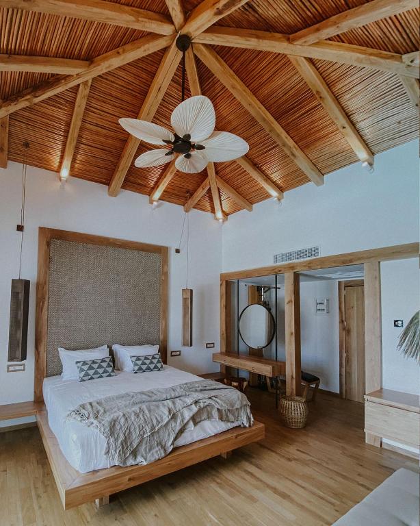 Mūsdienīgs guļamistabas interjers ar koka griestiem un elegantu ventilatoru, kas radīts relaksējošai atmosfērai. Gultā ir dekoratīvi spilveni, un blakus ir stilīgs spogulis un koka galds.